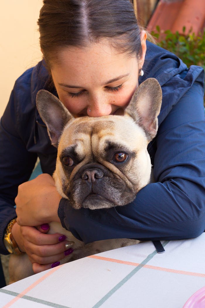 From above of crop smiling female with closed eyes embracing cute purebred dog while sitting at table outdoors